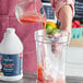 A hand pouring Capora Strawberry Banana Smoothie Mix into a blender with ice.