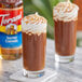 A glass of brown liquid with whipped cream on top next to a bottle of Torani Salted Caramel Flavoring Syrup.