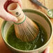 A hand using a Tenzo bamboo whisk to stir green liquid in a bowl.