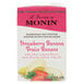 A carton of Monin strawberry banana fruit smoothie mix with a banana and strawberries on a plate.