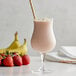 A pink strawberry banana smoothie in a glass with a yellow and white striped straw, with a strawberry and banana on the table.