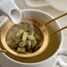 A white pitcher pouring liquid over green cardamom seeds in a gold sieve.