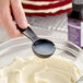 A black measuring spoon filled with liquid being held over a bowl of cream cheese mixture.
