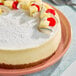 A round cheesecake topped with powdered sugar and decorated with pineapple, cherries, and coconut pieces on a pink plate.