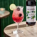 A bottle of Monin Premium Blackberry Flavoring Syrup next to a glass of iced blackberry drink garnished with a lime slice and blackberries.