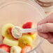 A hand holds a measuring spoon with Add A Scoop Fiber Blend supplement powder over a bowl of fruit.