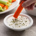 A person dipping a carrot into a bowl of white sauce.
