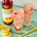 Two glasses of pink raspberry lemonade with ice and berries next to a Monin Sugar Free Raspberry Fruit Syrup bottle.