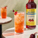 A bottle of Monin Sugar-Free Raspberry Flavoring Syrup next to a glass of iced raspberry beverage garnished with berries and mint.
