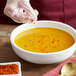 A person's gloved hand pouring Regal Extra Hot Ground Cayenne Pepper into a bowl of soup.