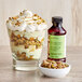 A dessert in a glass with a bowl of nuts and a bottle of LorAnn Oils Pistachio Emulsion.