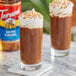 A glass of brown liquid with whipped cream on top next to a Torani Salted Caramel Flavoring Syrup bottle.