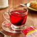 A glass cup of Bigelow Red Raspberry tea with a tea bag on a saucer.