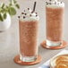 Two glasses of brown Frozen Bean Java Chip blended ice coffee with whipped cream and chocolate chips on a table.