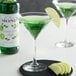 A glass of green liquid with a slice of green apple on the rim, next to a bottle of Monin Premium Granny Smith Apple Syrup.
