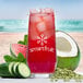 A glass of red Smartfruit Replenish beverage with ice and cucumber slices on the rim, next to a watermelon and coconut.