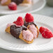 A plate with two chocolate-dipped cannoli filled with pink raspberry cream, topped with fresh raspberries and dusted with powdered sugar, accompanied by extra raspberries.