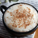 A cup of coffee with foam and brown sprinkles next to a black container of Regal Ground Nutmeg.