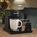 A Hamilton Beach black single serving pod coffee maker on a table with a white mug and packets.