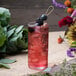 A glass of red liquid with berries and ice on a table with a Monin Blackberry Concentrated Flavor bottle.