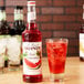 A bottle of Monin watermelon syrup next to a glass of watermelon soda with ice.