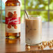 A glass of brown liquid with a straw and Torani Puremade Hazelnut Syrup on a table with nuts.