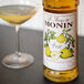 A bottle of Monin pear syrup next to a glass of yellow liquid.