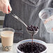 A person using a stainless steel wire scoop to add black tapioca balls to a clear plastic cup.