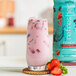 A glass of pink Smartfruit Renew Strawberry Acai drink next to a bottle of Smartfruit Renew Strawberry Acai concentrate.