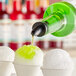 A bottle of green syrup being poured onto a cup of shaved ice, with two additional cups of plain shaved ice nearby.