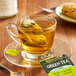 A glass cup of Bigelow Green Tea with Mint on a saucer with a tea bag in it on a table with a plate of cookies.