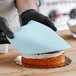 A person wearing black gloves using a knife to cut a blue cake with Satin Ice Baby Blue Vanilla Rolled Fondant on it.