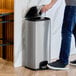 A man using a Lancaster Table & Seating rectangular decorative step-on trash can to throw away a bottle.