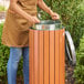 A round outdoor trash can with a wood composite exterior and open steel lid, being used by a person.
