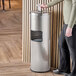 A woman standing next to a Lancaster Table & Seating stainless steel trash can with a sanitizing wipe dispenser.