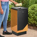 A square decorative trash can made of steel and wood with a modern design, placed outdoors.