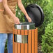 A round outdoor trash can with a wood composite exterior, steel inner bin, and a side opening lid.