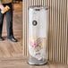 A person standing next to a Lancaster Table & Seating clear plastic decorative trash can with a stainless steel top.