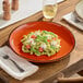 A plate of pasta with tomatoes and basil on an Acopa Keystone Sedona Orange porcelain coupe plate on a wooden table.