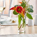 An Acopa cylindrical glass bud vase with flowers and leaves on a table.