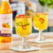 A glass of yellow mango-flavored beverage with ice, fruit slices, and a chili powder rim, next to a bottle of Capora Mango Flavoring Syrup.