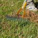 A metal thatch rake with a yellow adjustable head and a hardwood handle being used to remove debris from grass.