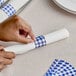 A hand using blue and white checkered self-adhering paper napkin band to wrap a napkin.