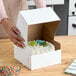 A person opening a Baker's Lane white cake box with a cake inside.