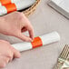 A person's hands using an orange and white Choice paper napkin band to wrap a napkin around silverware.