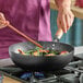 A 12-inch carbon steel non-stick flat bottom wok with a wood handle being used to cook vegetables and shrimp on a stovetop.