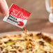 A hand holding a Pizza Packet Crushed Red Pepper packet over a pizza.