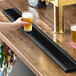 A long, black matte stainless steel surface-mount beer drip tray on a wooden bar counter.