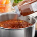 A person pouring McCormick Culinary Light Chili Powder into a pot of chili on a stove.