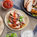 A tortilla with meat and vegetables on it with red and white sauce on the side. A table with sliced cucumbers and other ingredients.